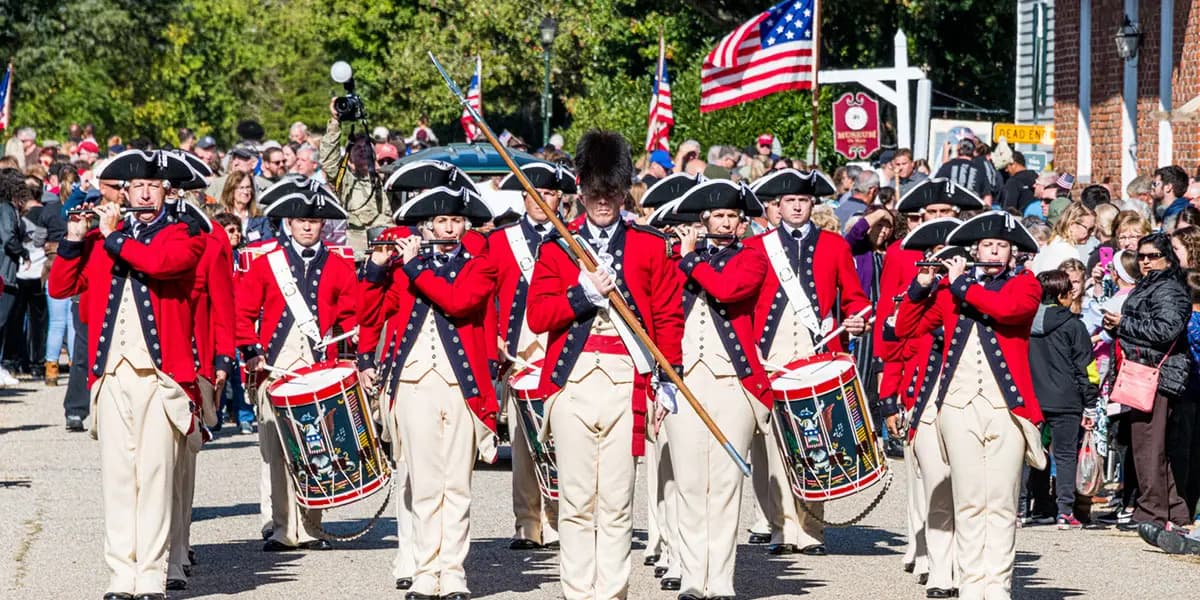 A group of musicians in Revolutionary War-era uniforms march in a parade, playing instruments and carrying drums.