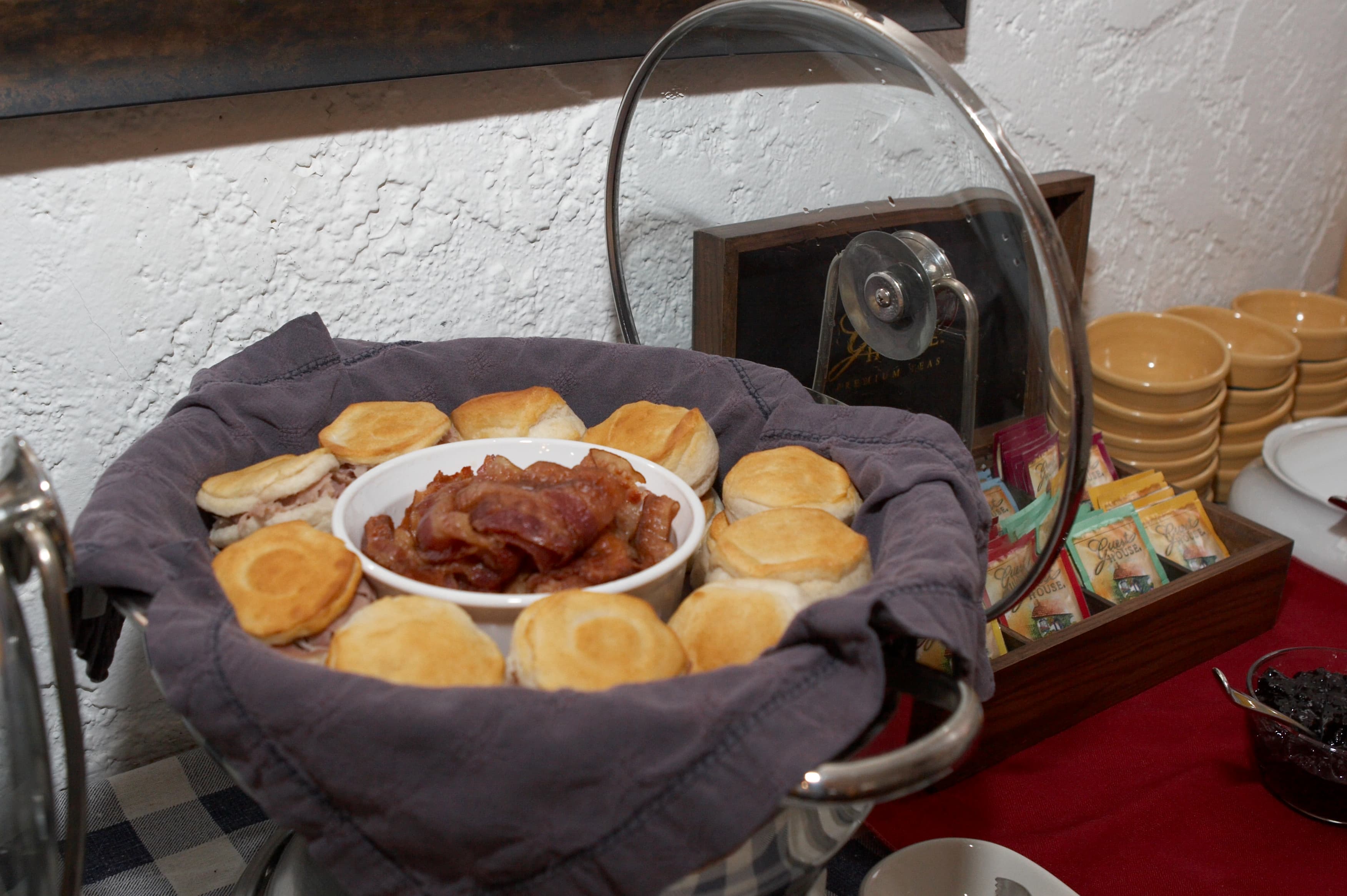 Bacon and biscuits in a metal serving bowl with hinged clear lid with tea in packets and yellow bowls in the background