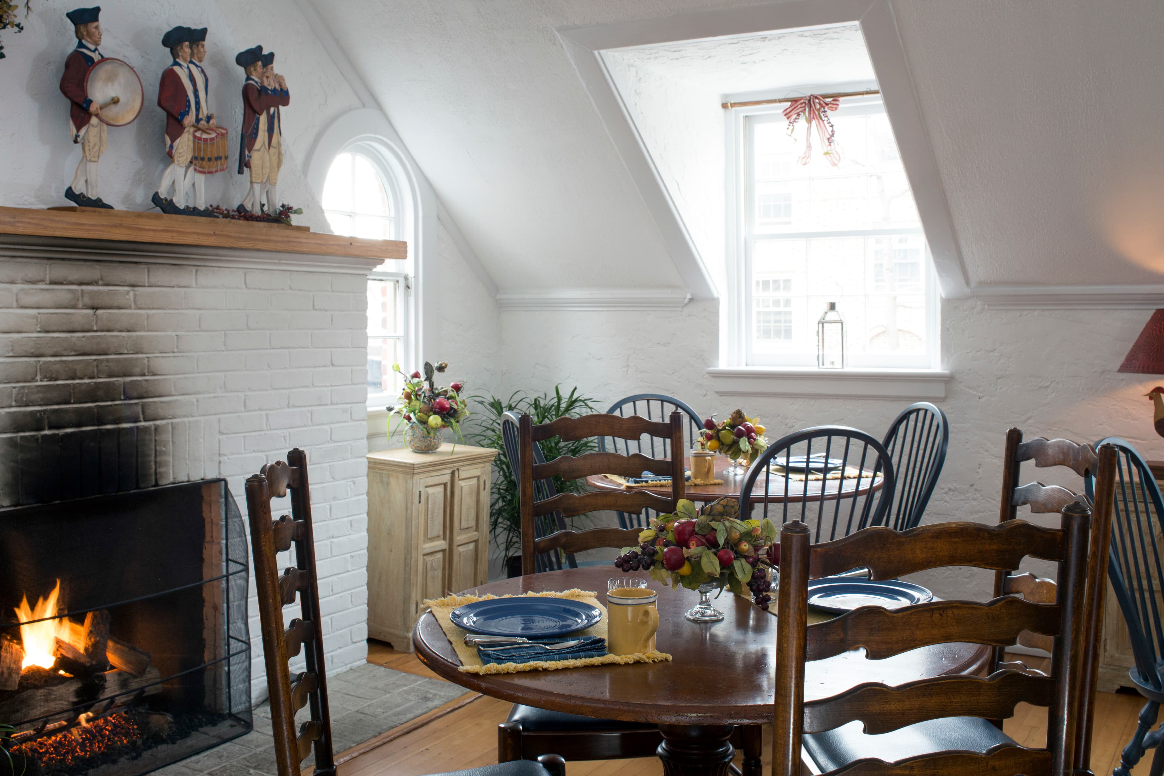 A dining room with several small tables with tablecloths and wood chairs, and a fire going in a fireplace in a white brick hearth.