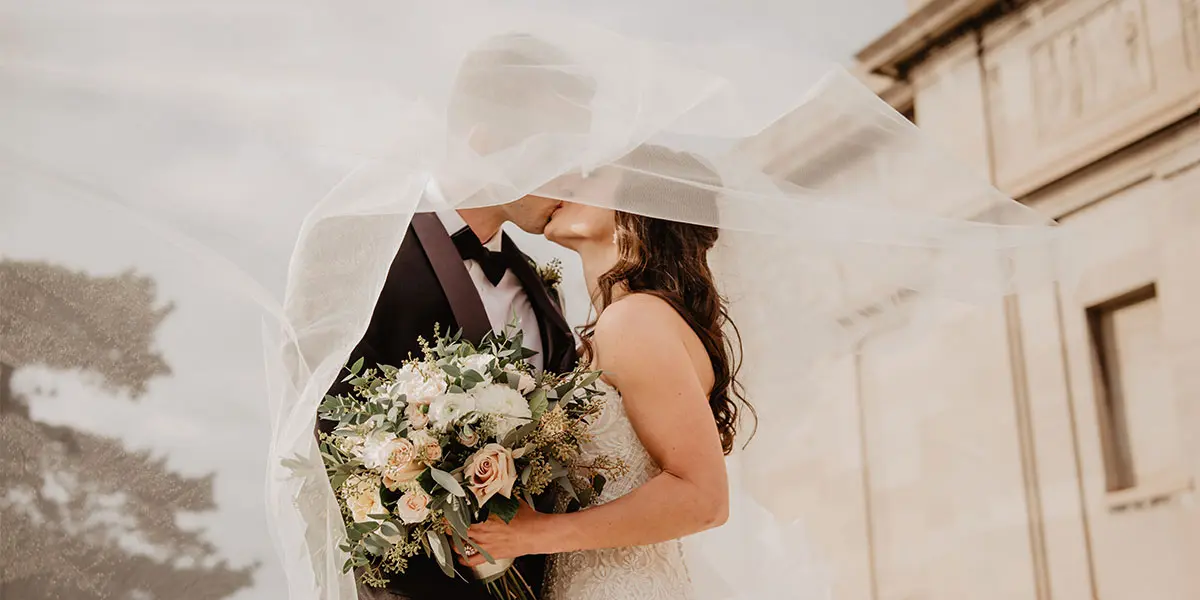 A couple kisses under a flowing veil, holding a bouquet of flowers.