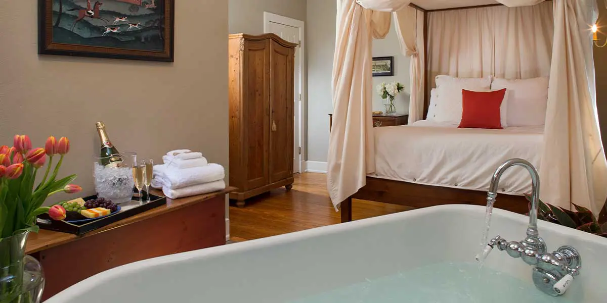 A cozy hotel room featuring a four-poster bed, a bathtub with running water, and a small refreshment station.