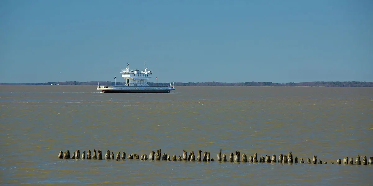 A ferry navigates across a calm, brownish body of water with a line of wooden pylons in the foreground.