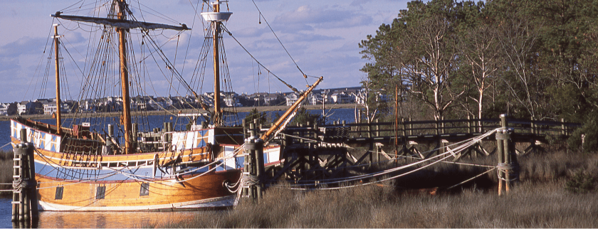 A wooden ship docked at a pier surrounded by greenery and water.
