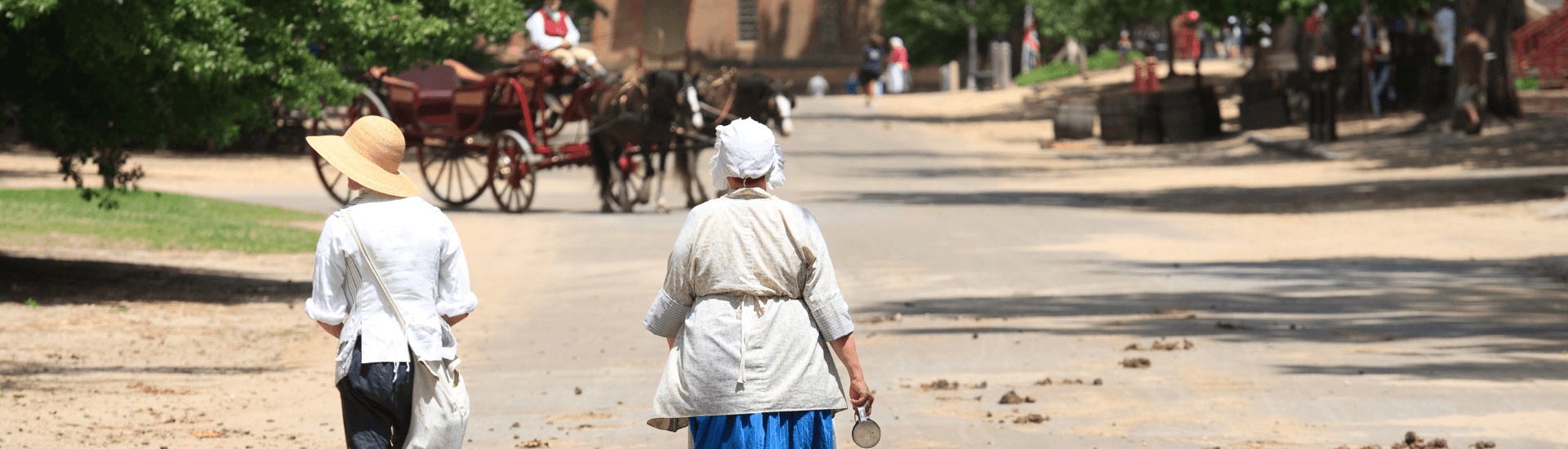 A man and woman dressed in 18th century clothing walking down a dirt street with a horse and buggy coming towards them.