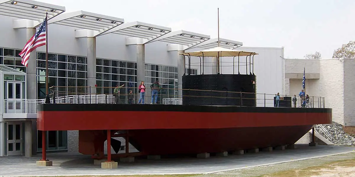 A large, red ship replica stands outside a modern building, with visitors exploring the deck.