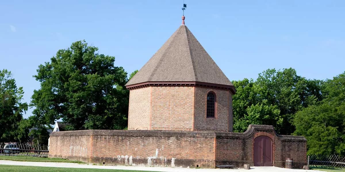 A brick round building with a conical roof, surrounded by trees and a wall.