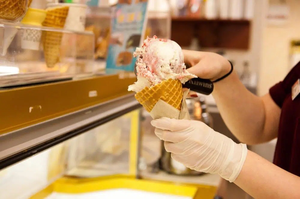 A server scoops ice cream into a waffle cone while wearing gloves.
