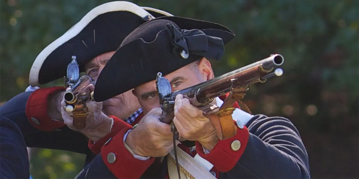 Two men in historical military attire aim flintlock rifles.